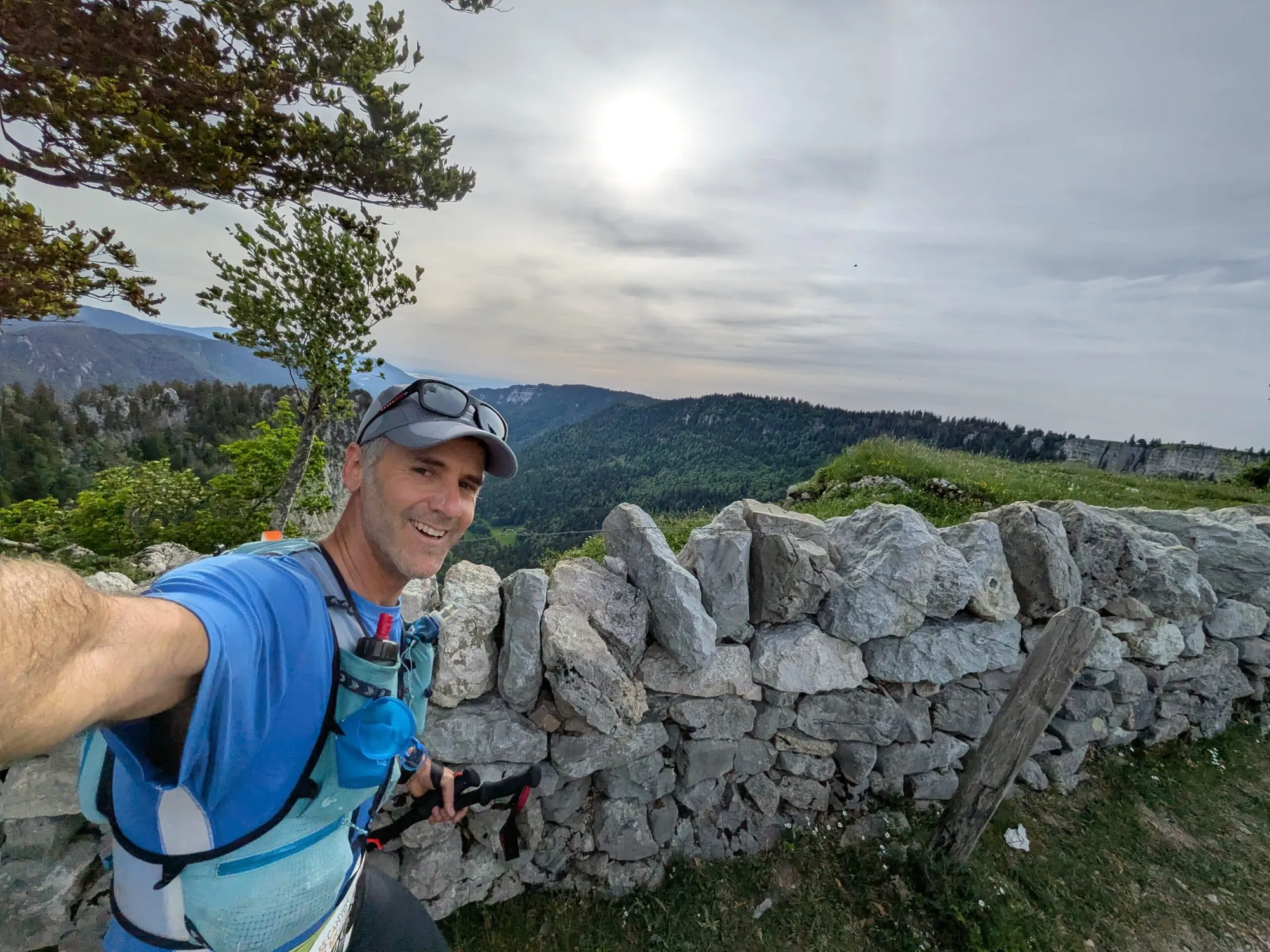 Smiling man in a blue hiking shirt and gray cap takes a selfie against a stone wall with mountains and a cloudy sky behind him.