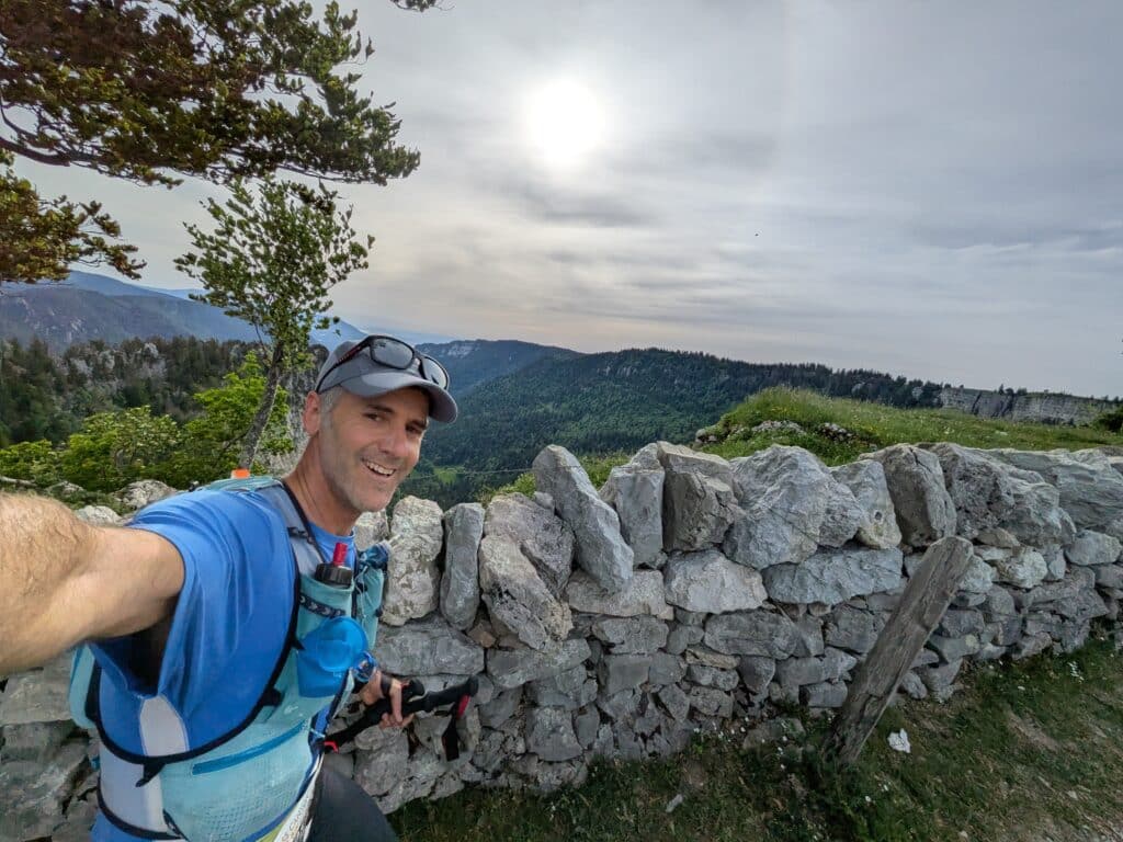 Smiling man in a blue hiking shirt and gray cap takes a selfie against a stone wall with mountains and a cloudy sky behind him.